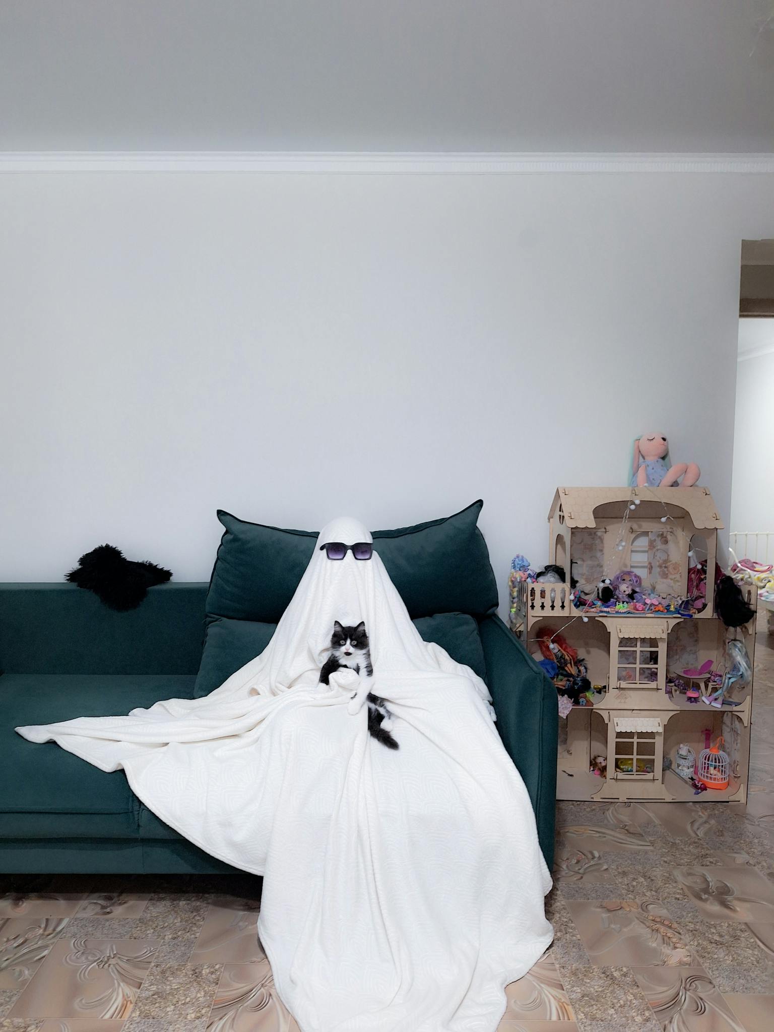 A person in ghost costume sits on a couch holding a black cat, with a dollhouse nearby.