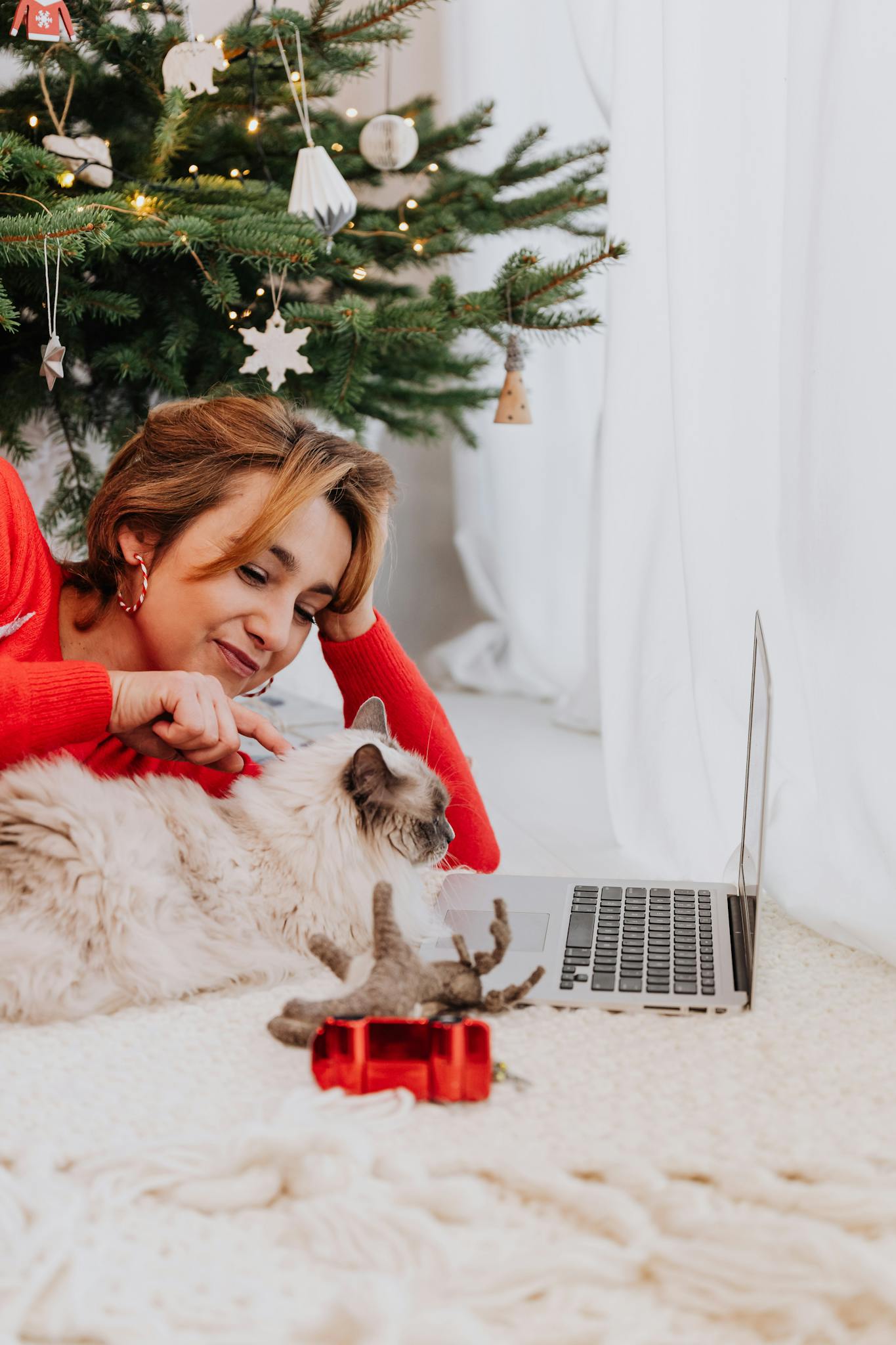 Happy woman petting her cat under a Christmas tree with a laptop nearby, celebrating the holidays.