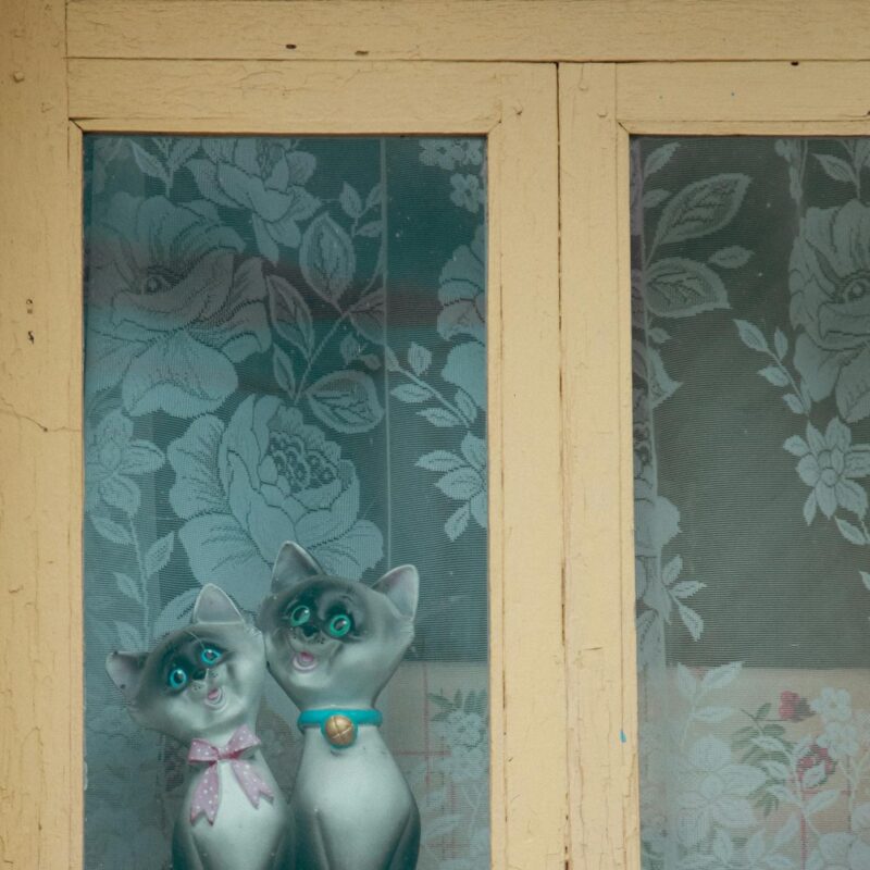 Two adorable cat figurines sit behind a vintage window with floral curtains.